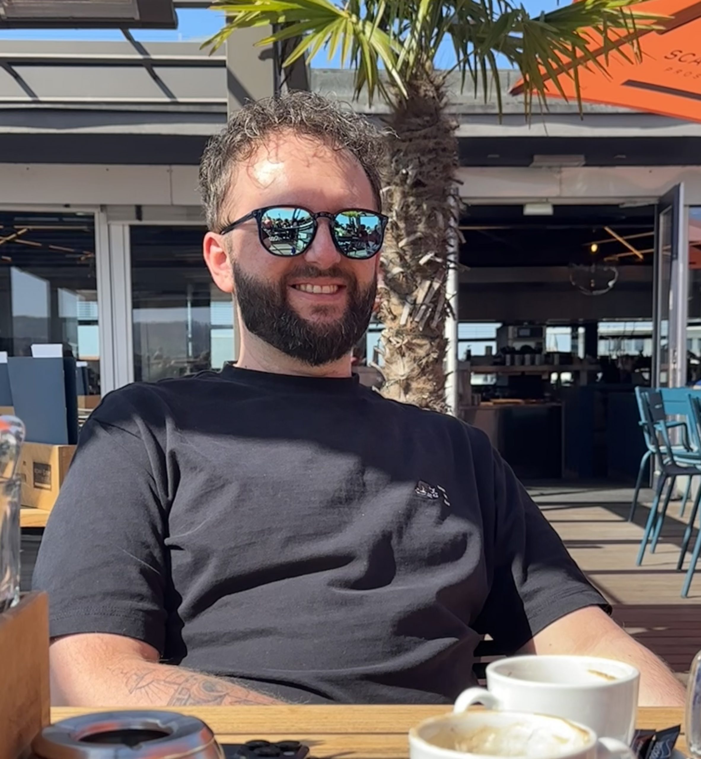 Mann mit Sonnenbrille sitzt entspannt im Freien in einem Café, vor sich Kaffeetassen, im Hintergrund Palmen und Sonnenschirm.